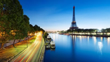 HD desktop wallpaper featuring the illuminated Eiffel Tower at night beside the Seine River, with light trails from passing vehicles along a tree-lined road.