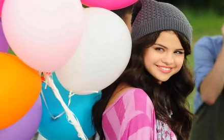 HD desktop wallpaper of Selena Gomez holding colorful balloons, wearing a gray beanie and pink top, smiling in an outdoor setting. Tags include music, Selena Gomez.