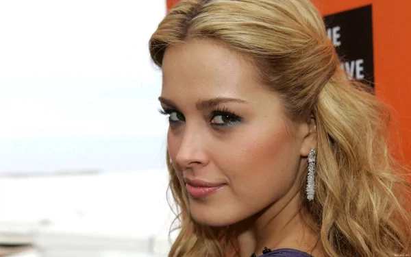 Close-up HD desktop wallpaper of a blonde female celebrity glancing over her shoulder, wearing dangling earrings and subtle makeup against a soft, neutral background.