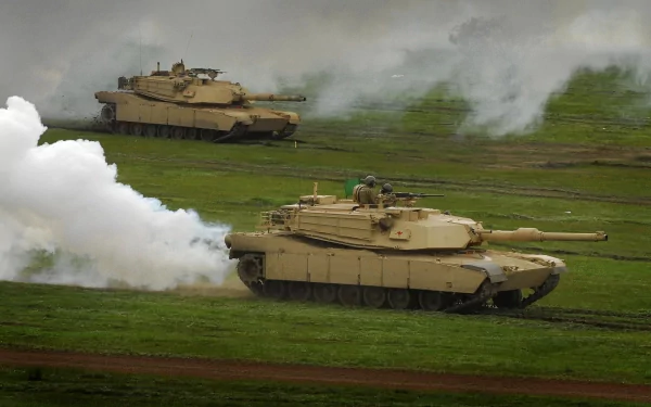 HD desktop wallpaper showing two military M1 Abrams tanks advancing across a grassy field with smoke surrounding them.