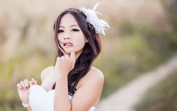 Asian woman with long dark hair, wearing a white dress and a feather hair accessory, stands in a field. HD desktop wallpaper and background.