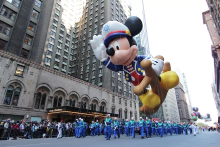HD desktop wallpaper capturing a vibrant parade scene with a large Mickey Mouse balloon floating above a marching band amidst tall city buildings.