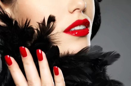 A close-up of a woman's lips adorned with vibrant red lipstick, complemented by striking red nails and a black feather accessory, set against a smooth gray background. 4K Ultra HD wallpaper.