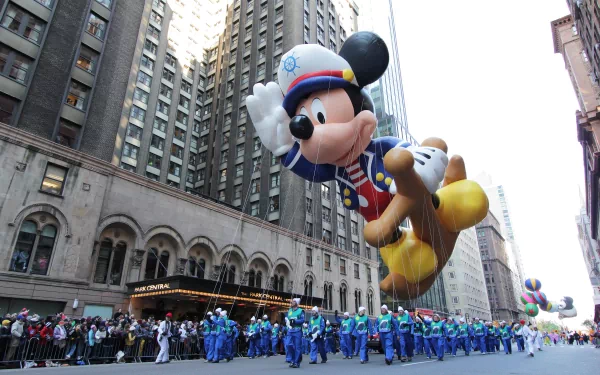 HD desktop wallpaper capturing a vibrant parade scene with a large Mickey Mouse balloon floating above a marching band amidst tall city buildings.