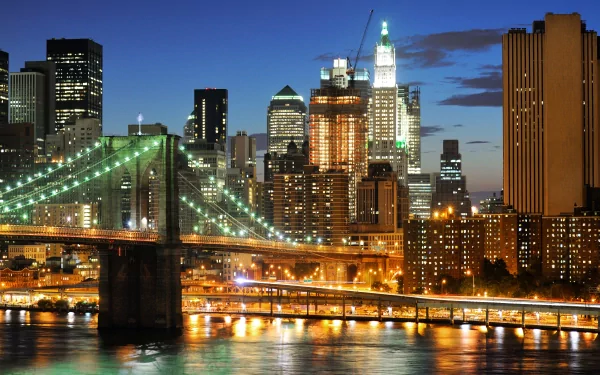 4K Ultra HD image of New York City's illuminated skyline at dusk, featuring the Brooklyn Bridge and iconic skyscrapers reflecting on the water.