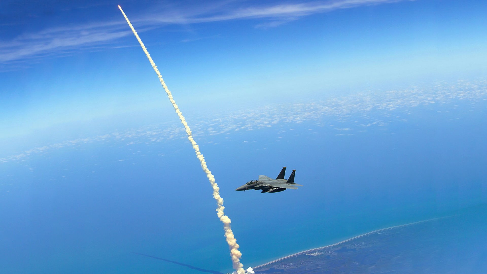 Military McDonnell Douglas F-15 Eagle jet flying over the ocean with a missile launch trail against a clear blue sky, captured as an HD PC desktop wallpaper background.