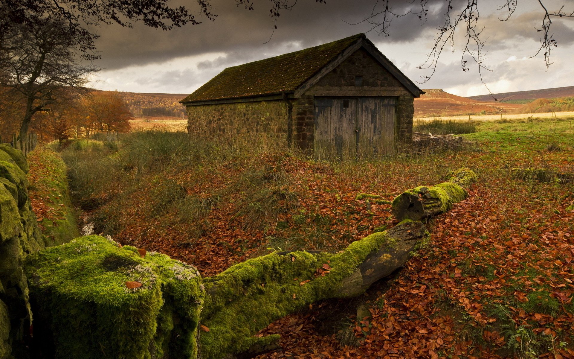 HD PC desktop wallpaper featuring a rustic man-made house in an autumn landscape with moss-covered rocks and overcast skies.