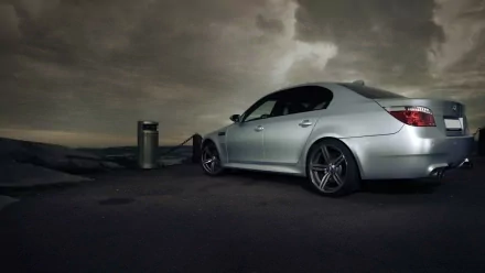 HD desktop wallpaper featuring a silver BMW E60 parked under a dramatic, cloudy sky at dusk.