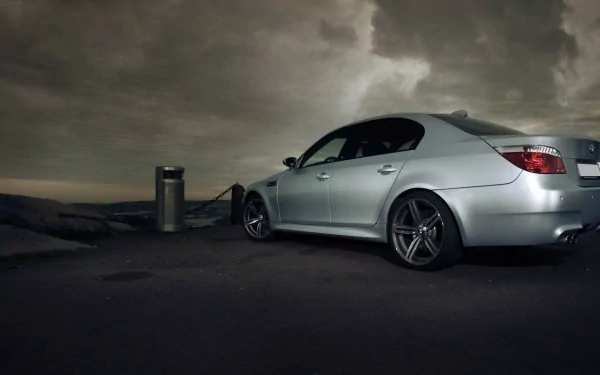 HD desktop wallpaper featuring a silver BMW E60 parked under a dramatic, cloudy sky at dusk.