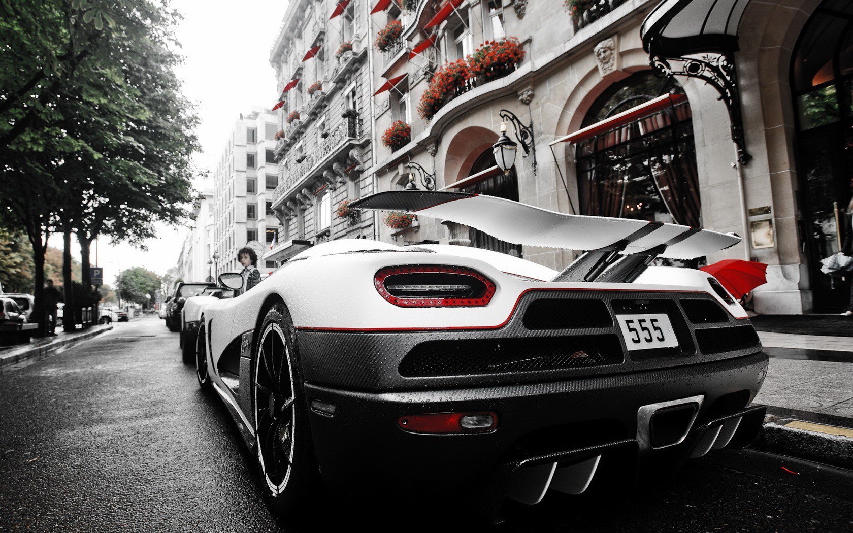 HD PC desktop wallpaper: rear view of a white Koenigsegg vehicle on a wet city street, carbon accents and red trim visible, ornate buildings and flower boxes lining the sidewalk in the background.
