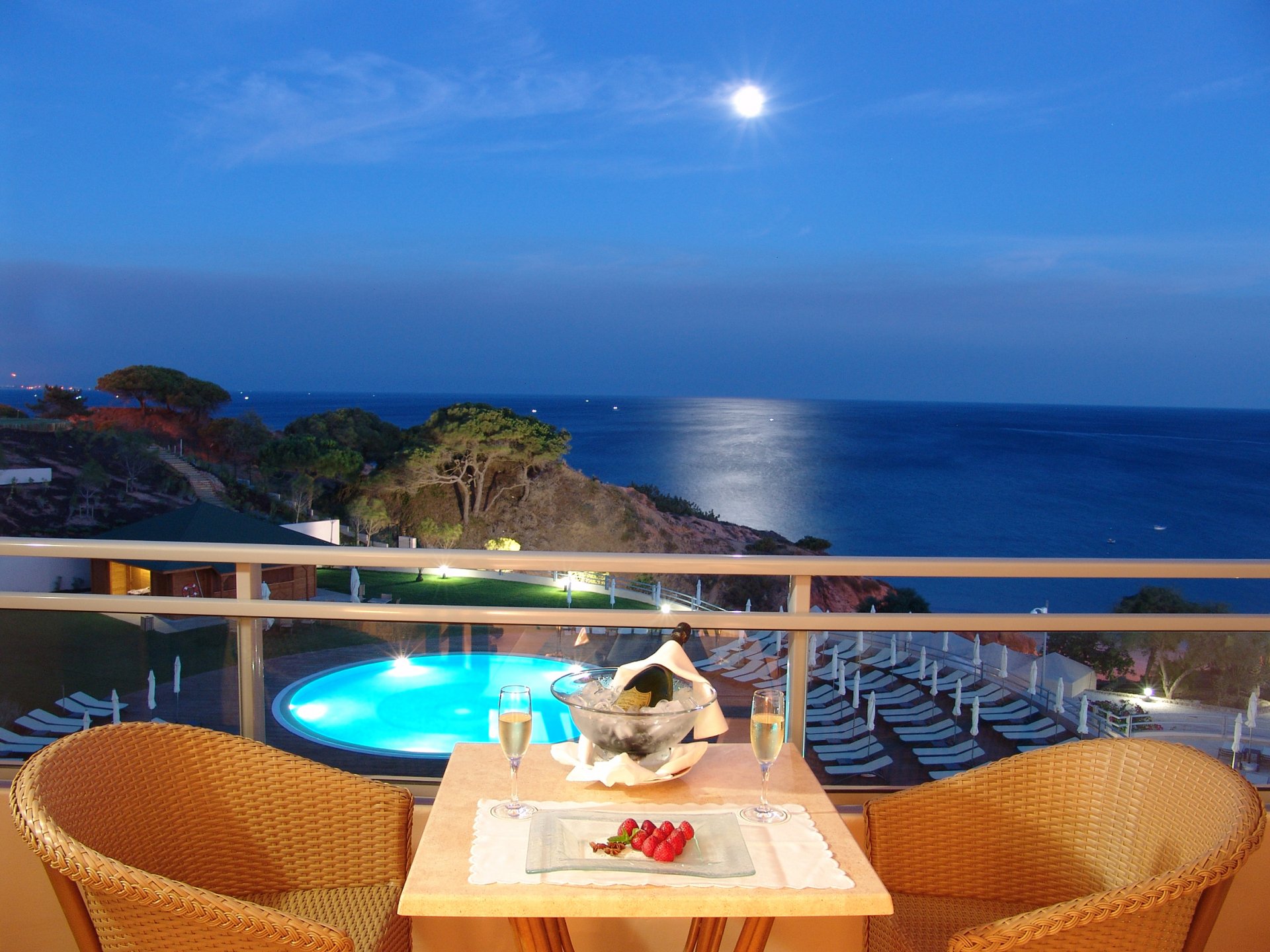 HD PC desktop wallpaper/background: man-made balcony with table, chairs and wine glasses overlooking a lit pool, moonlit sea and rocky coast at dusk.