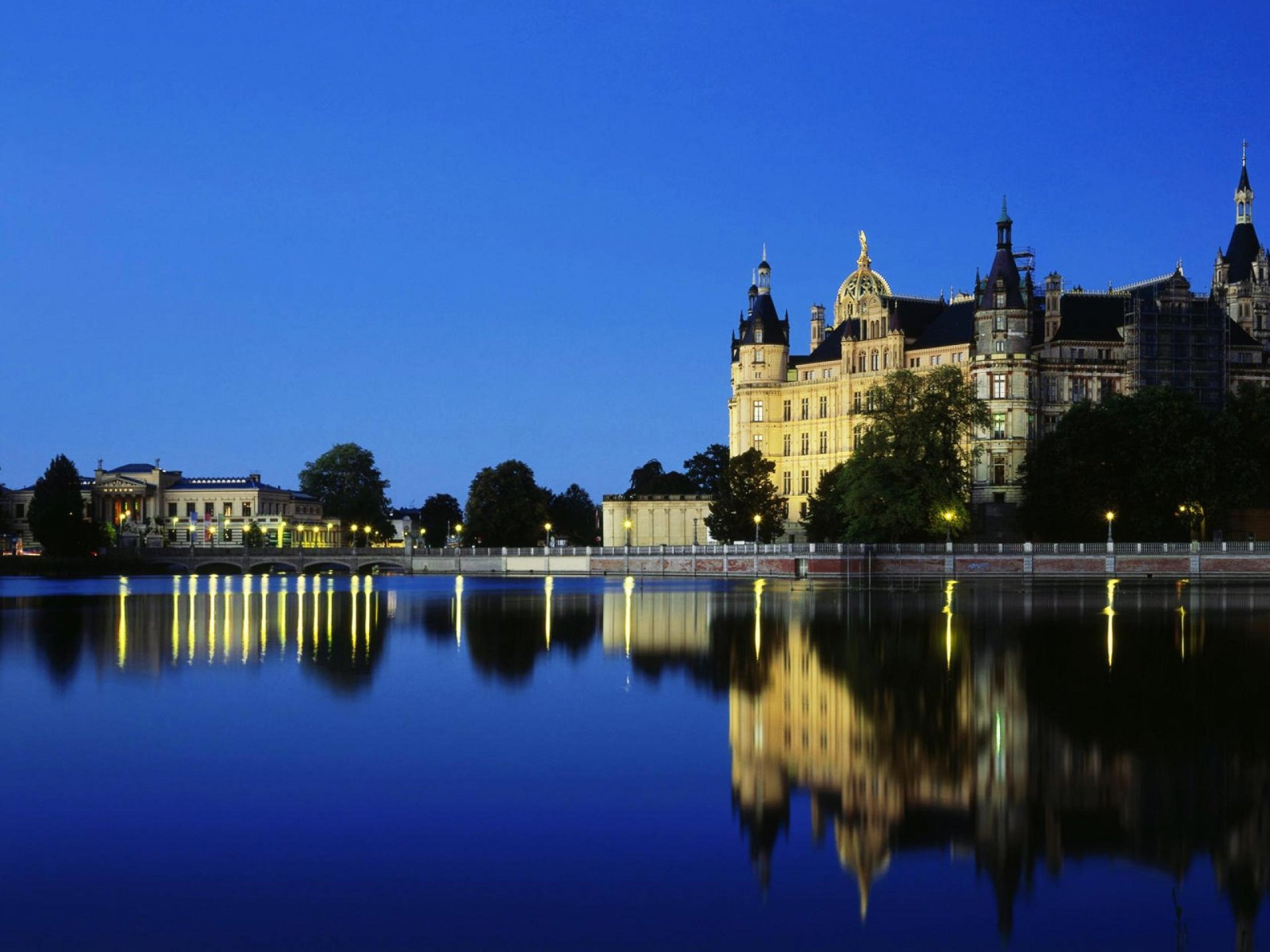 Man-made Schwerin Palace at twilight, its illuminated facade mirrored in a calm lake under a deep blue sky — 2K Quad HD PC desktop wallpaper and background.