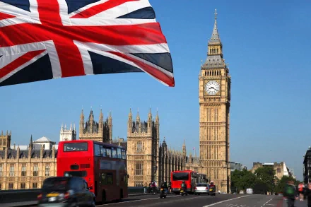 A vibrant view of Big Ben against a clear blue sky, with a Union Jack flag waving and red double-decker buses passing by, showcasing a lively scene of London in 4K Ultra HD.