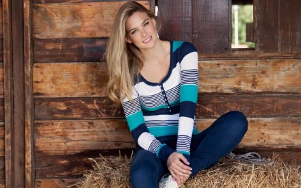 HD desktop wallpaper featuring celebrity Bar Refaeli sitting on hay inside a rustic wooden barn, dressed in a striped hoodie and jeans, smiling at the camera.