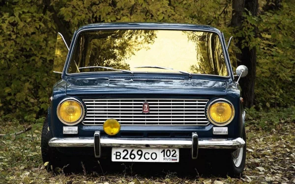 Front view of a blue VAZ-2101 vehicle with yellow headlights parked on a leaf-covered ground, set against a backdrop of dense green foliage, captured in HD for a PC desktop wallpaper.