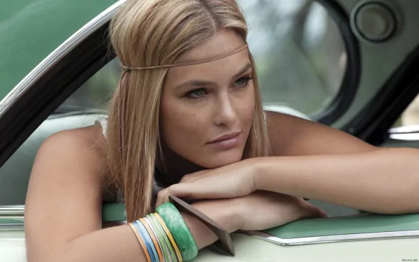 A close-up of Bar Refaeli resting her arms on a car window, showcasing her relaxed expression and stylish accessories in a natural, outdoor setting. High-definition desktop wallpaper.