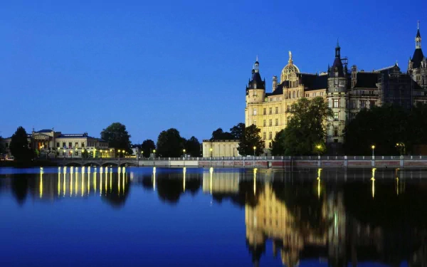 Man-made Schwerin Palace at twilight, its illuminated facade mirrored in a calm lake under a deep blue sky — 2K Quad HD PC desktop wallpaper and background.