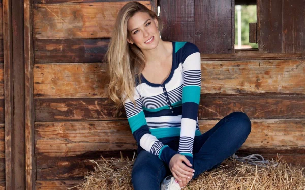 HD desktop wallpaper featuring celebrity Bar Refaeli sitting on hay inside a rustic wooden barn, dressed in a striped hoodie and jeans, smiling at the camera.