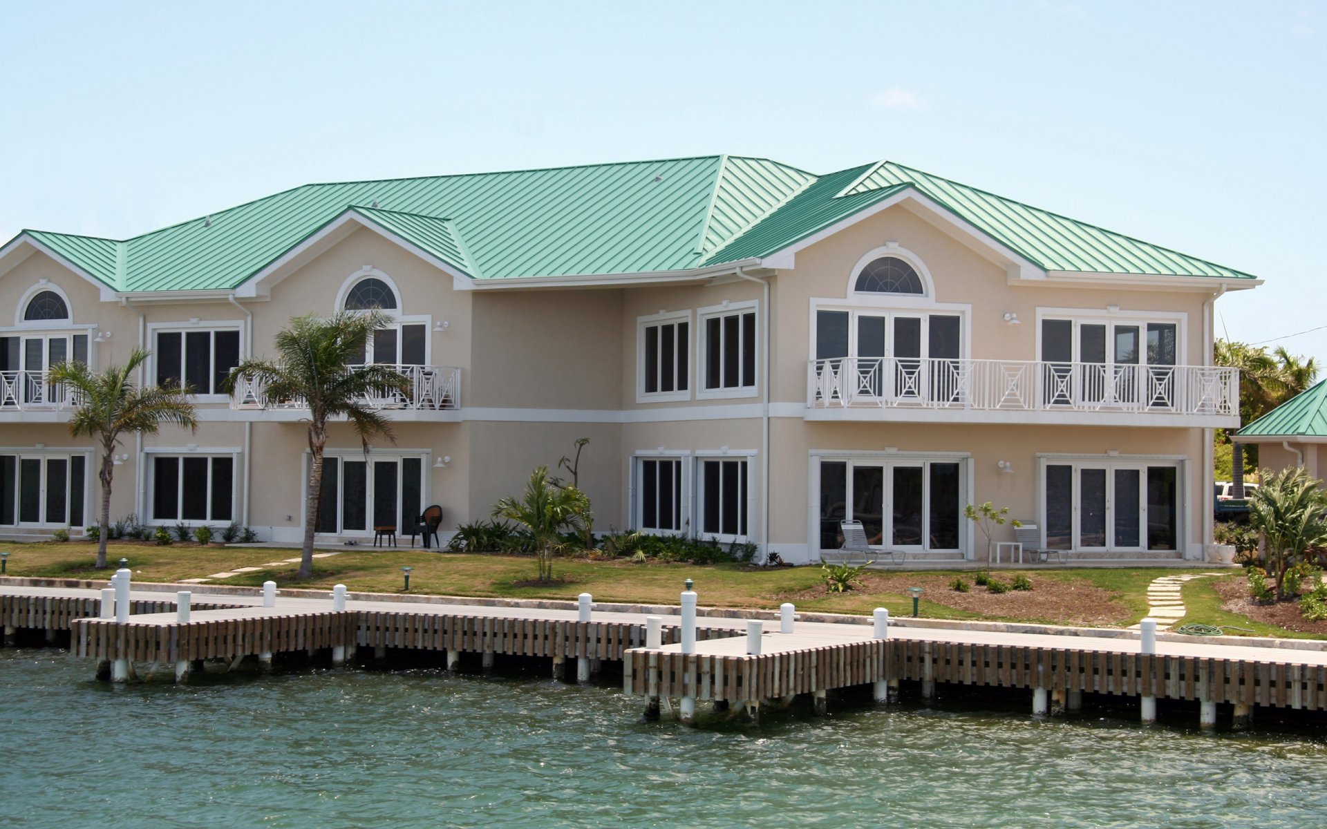 HD desktop wallpaper of a large man-made mansion with a green roof by the waterfront, featuring balconies and palm trees under a clear sky.