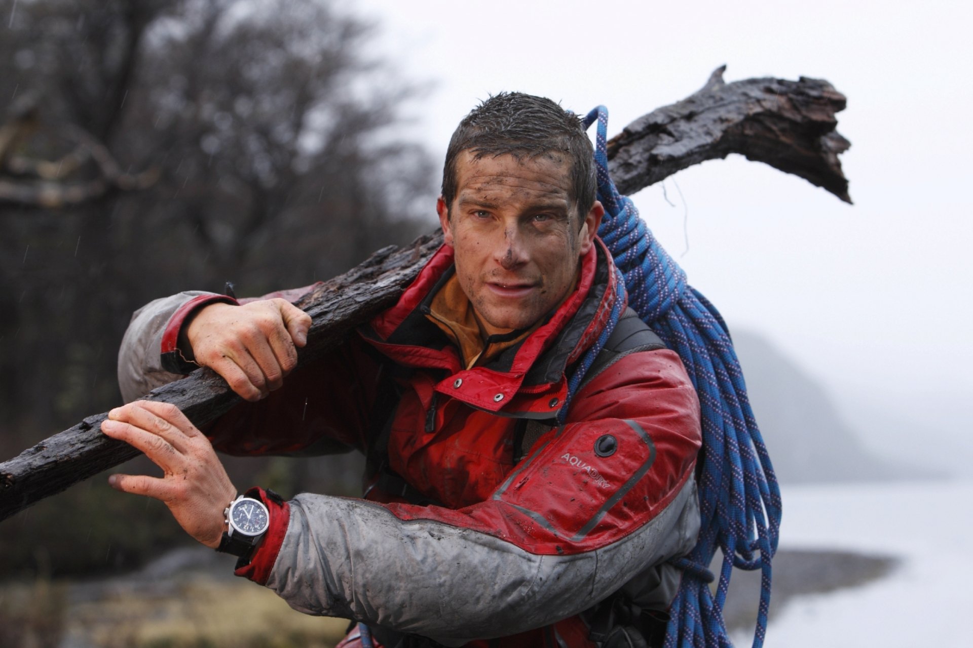 HD desktop wallpaper featuring Bear Grylls, dressed in outdoor gear, carrying a large stick with a coiled rope over his shoulder in a rugged, misty environment.