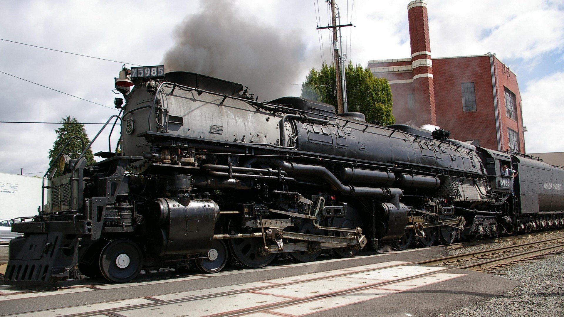 HD desktop wallpaper featuring a detailed black steam locomotive emitting smoke as it rests near industrial buildings and railroad tracks under a cloudy sky.