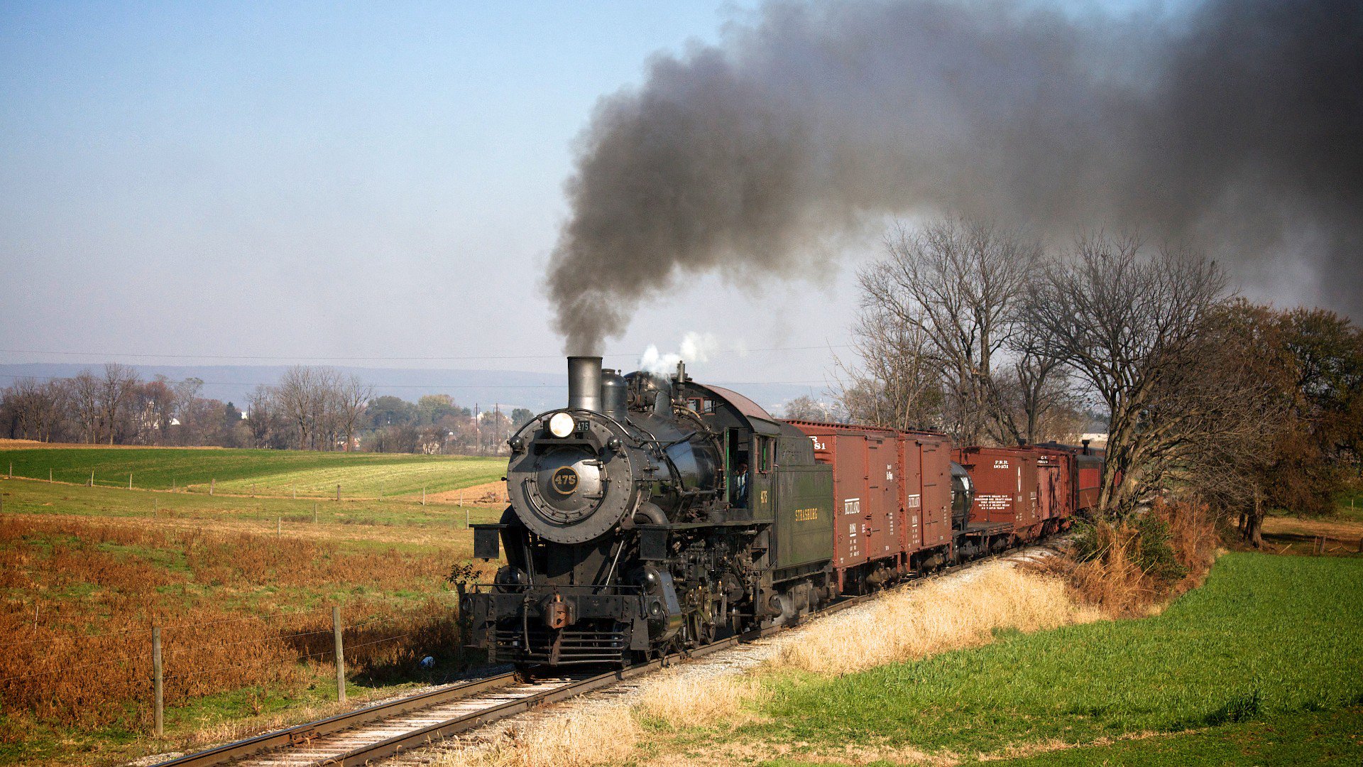 HD PC desktop wallpaper/background: vintage locomotive vehicle — a steam engine pulling freight cars across sunlit rural fields, trailing a plume of dark smoke.