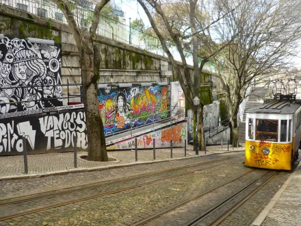 HD PC desktop wallpaper: a yellow tram vehicle on cobblestone tracks passing colorful graffiti on stone walls, trees and steps in an urban streetscape.