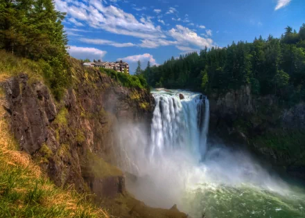 Snoqualmie Falls cascades powerfully down a cliff surrounded by lush forest, with a house perched near the edge and a stream flowing below under a bright blue sky.