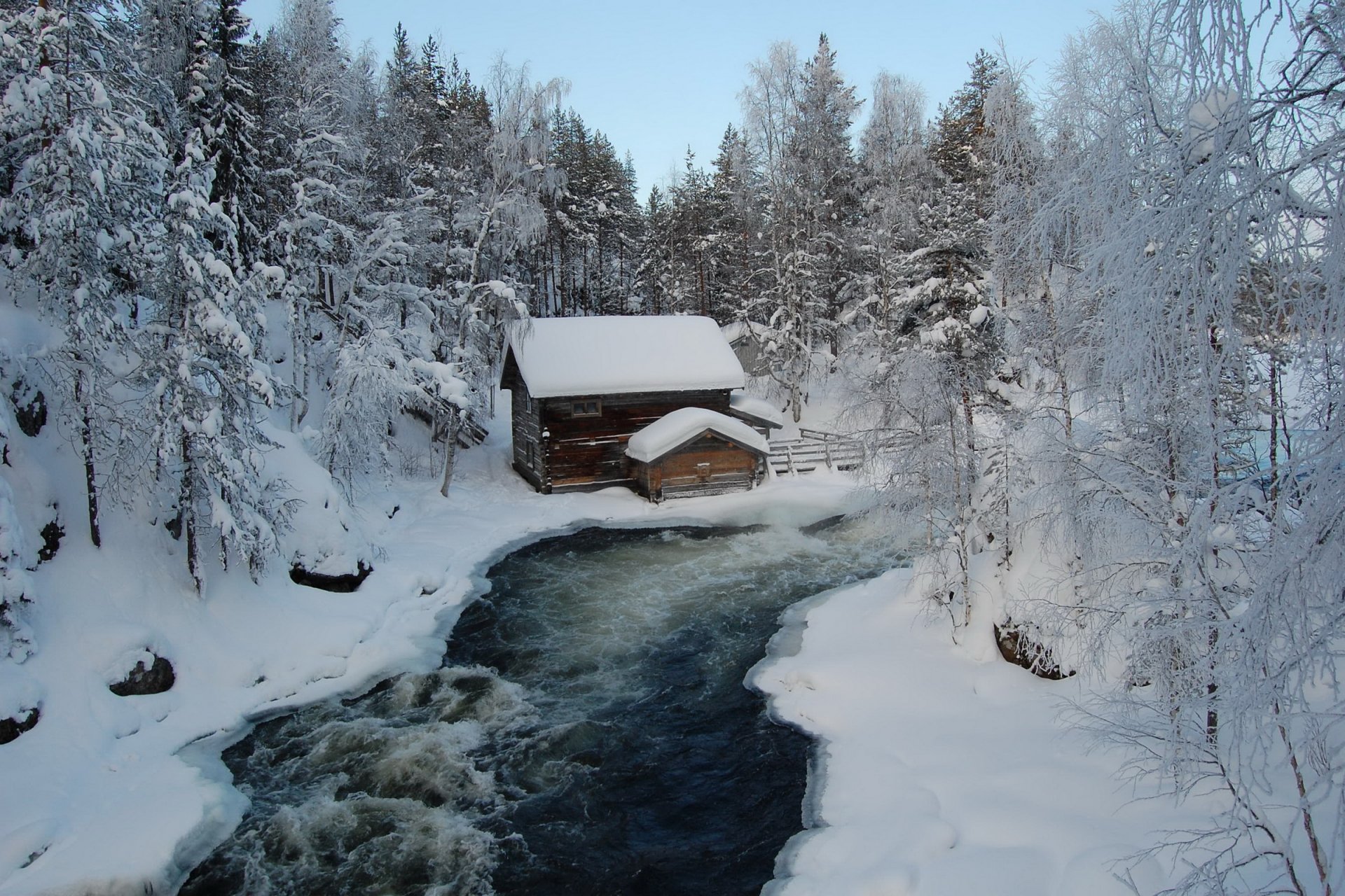 HD desktop wallpaper of a man-made cabin nestled in a snowy forest beside a flowing, partially frozen river.