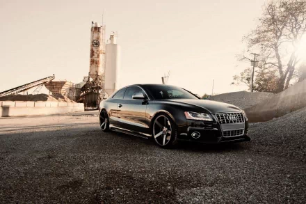HD PC desktop wallpaper featuring a sleek black Audi vehicle parked on a gravel surface with industrial structures and trees in the background.