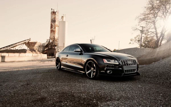 HD PC desktop wallpaper featuring a sleek black Audi vehicle parked on a gravel surface with industrial structures and trees in the background.