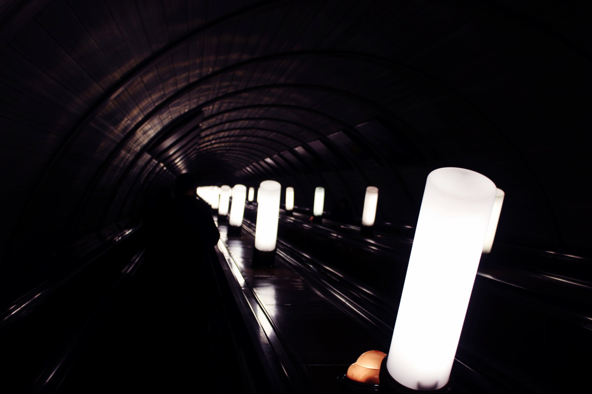 Dark underground man-made subway tunnel with glowing cylindrical lights receding into the distance, a hand holding a light in the foreground — 5K Ultra HD PC desktop wallpaper/background.