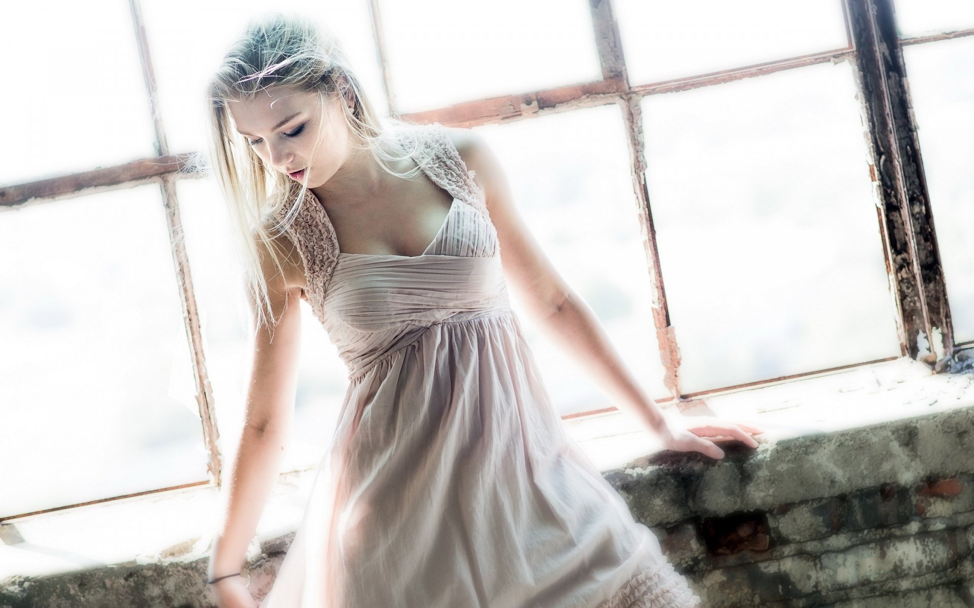 HD desktop wallpaper of a woman in a light dress standing by a large, rustic window, looking down contemplatively.