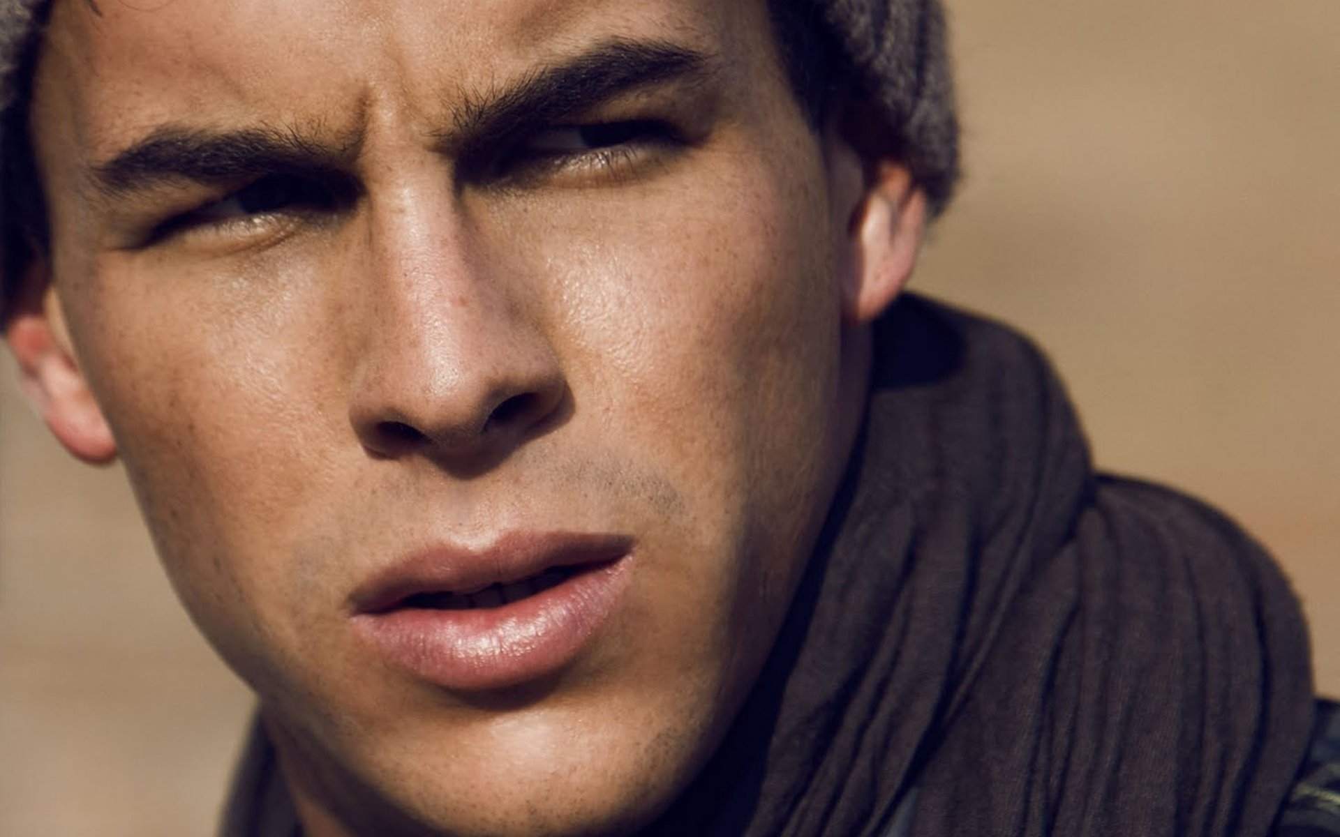 Close-up HD desktop wallpaper of celebrity Mario Casas wearing a beanie and scarf, with a warm-toned blurred background.