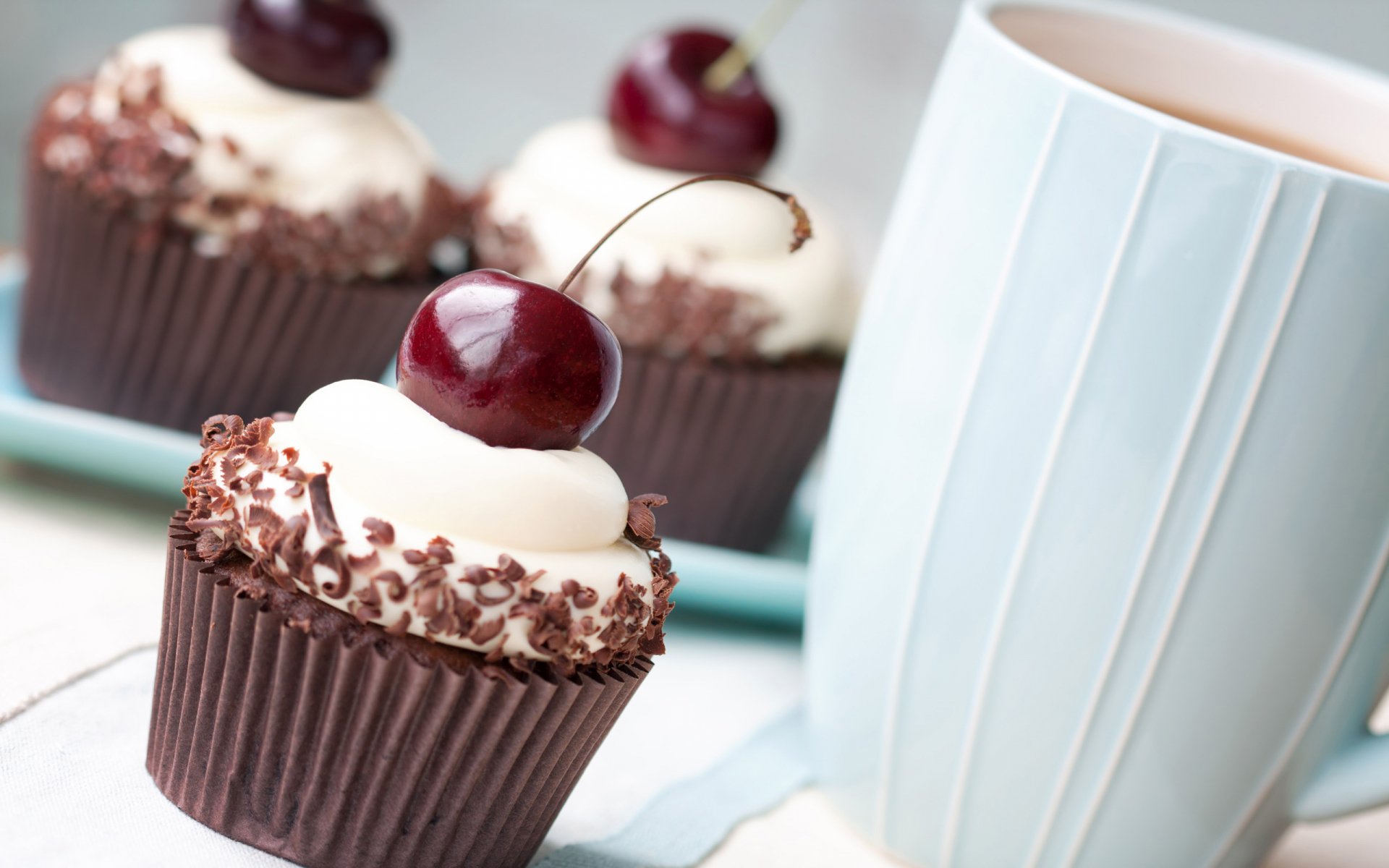 A close-up of delicious chocolate cupcakes topped with cream and cherries, alongside a light blue mug, creating an inviting food-themed HD desktop wallpaper.