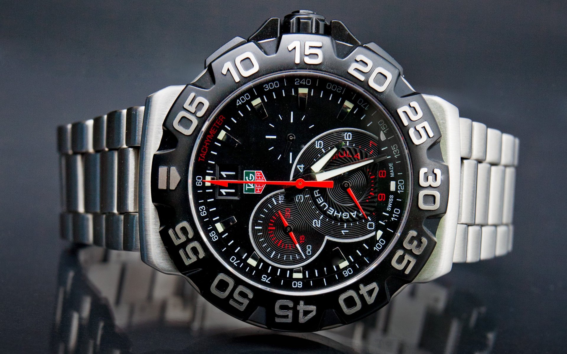 HD desktop wallpaper featuring a close-up of a man-made wristwatch with a detailed black dial and stainless steel band on a reflective surface.