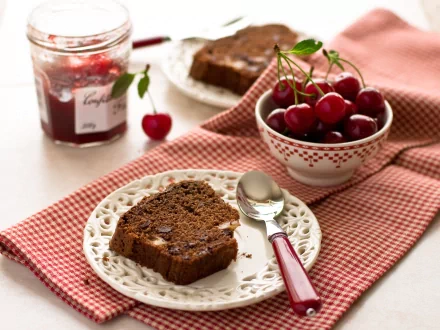 A delicious slice of chocolate cake sits on an ornate plate beside a bowl of fresh cherries, with a jar of cherry jam in the background, creating an inviting still life scene.
