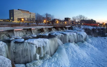 HD desktop wallpaper of Rideau Falls in Ottawa, Canada, featuring frozen cascading waters and city buildings under a clear evening sky.