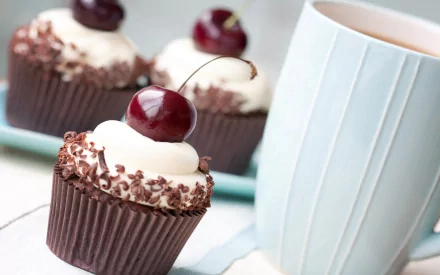 A close-up of delicious chocolate cupcakes topped with cream and cherries, alongside a light blue mug, creating an inviting food-themed HD desktop wallpaper.