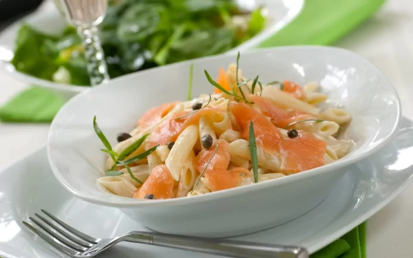 A close-up of a delicious pasta dish garnished with smoked salmon and herbs, set on a table with a green salad in the background. A vibrant HD wallpaper for food lovers.