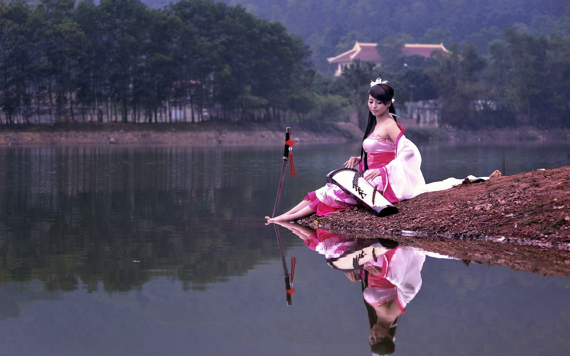 An Asian woman in traditional attire sits by a calm lake with a serene forest and temple in the background, creating a tranquil and picturesque HD desktop wallpaper.