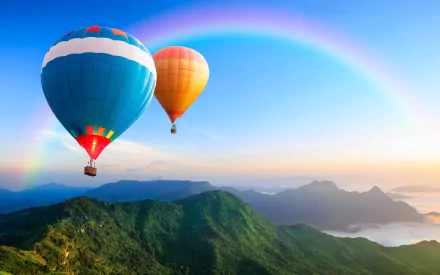 A vibrant HD wallpaper featuring two colorful hot air balloons soaring over lush mountains, with a rainbow arcing across a clear blue sky.