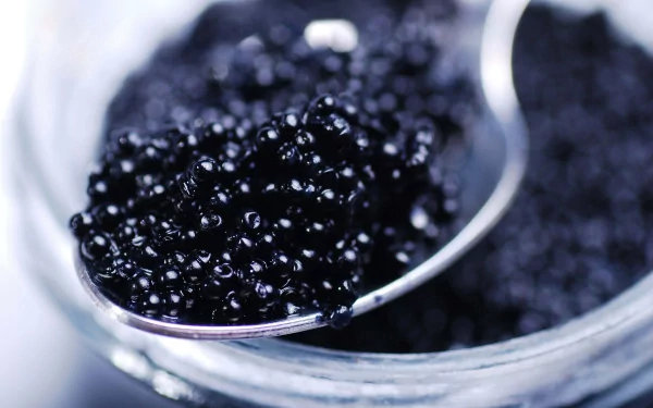 Close-up 4K Ultra HD desktop wallpaper of shiny black caviar served on a silver spoon above a glass jar.
