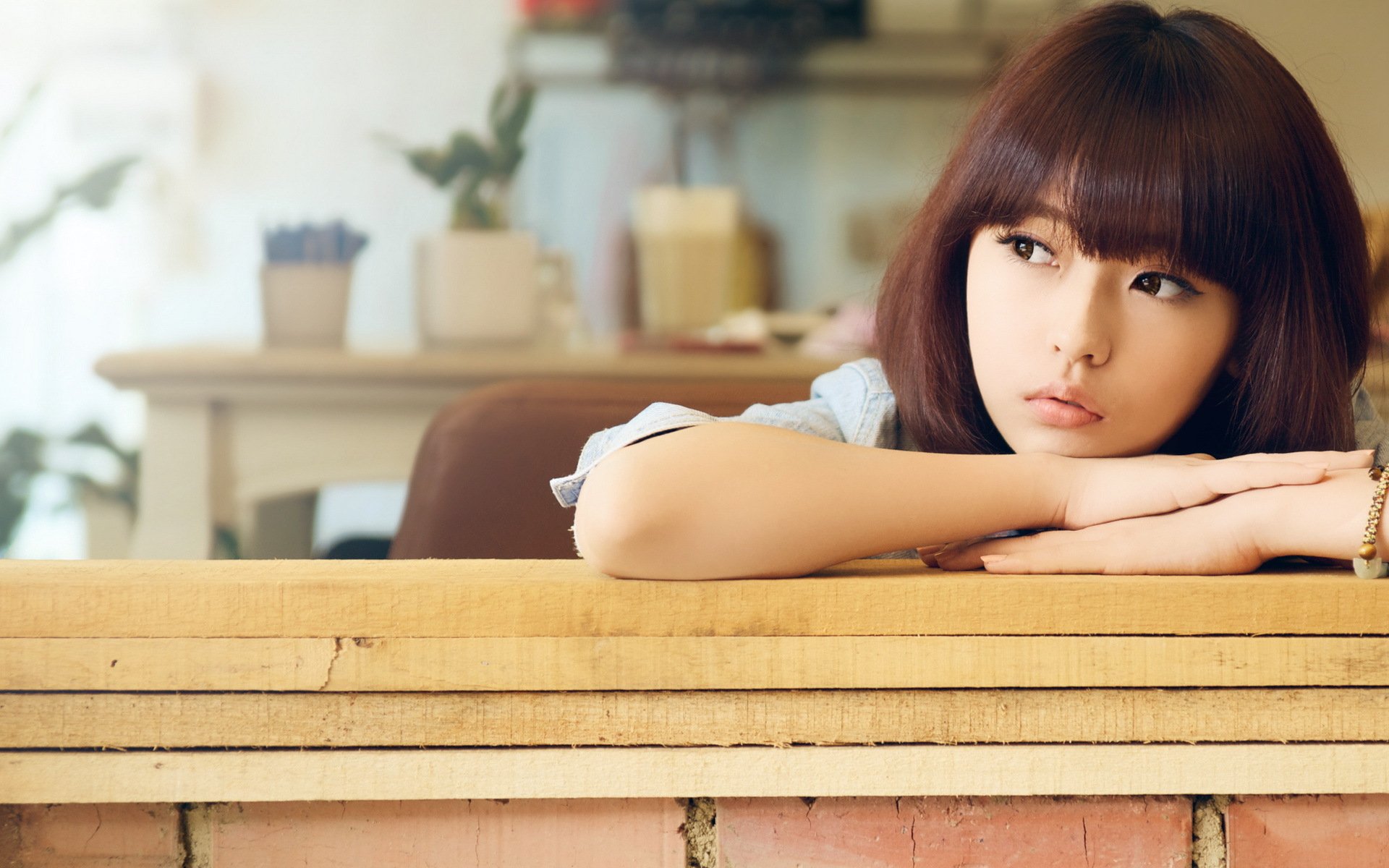 HD desktop wallpaper featuring an Asian woman with long dark hair, resting her chin on her arms while looking to the side in a thoughtful manner. The background has a cozy, indoor atmosphere with soft lighting.