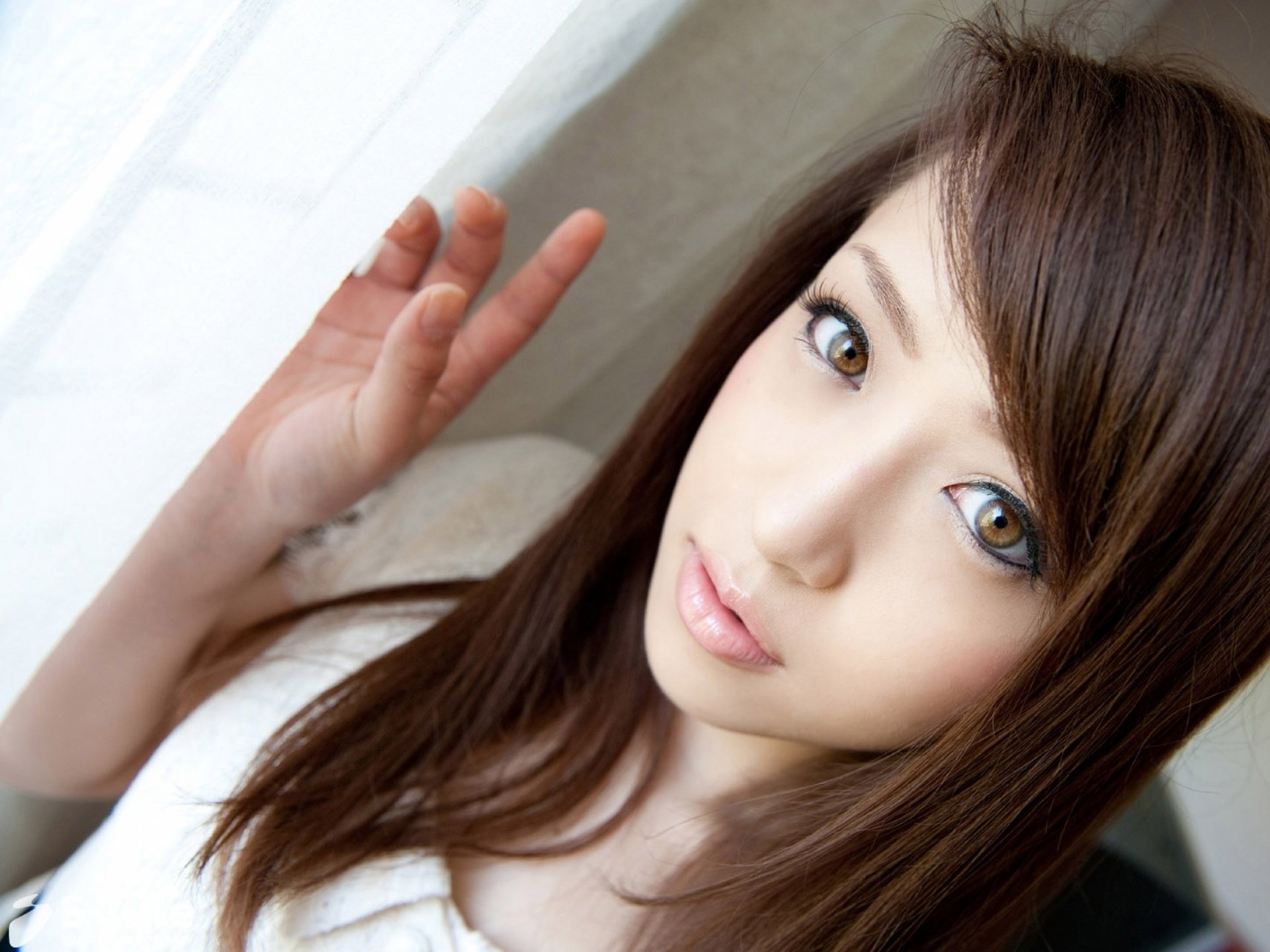 A close-up of a woman with long, brown hair and striking eyes, positioned near a window, making a peace sign. This HD image serves as an attractive desktop wallpaper and background.