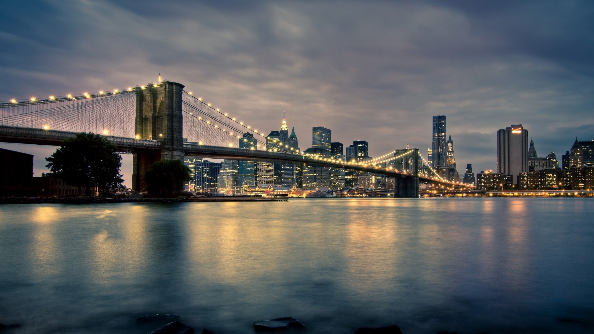 HD desktop wallpaper featuring the illuminated Brooklyn Bridge spanning over water with the Manhattan skyline in New York City under a moody evening sky.