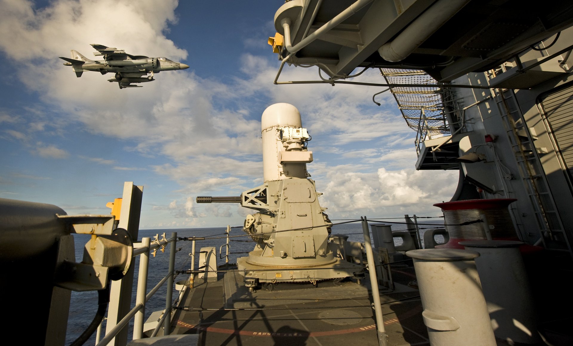 View from the deck of the USS Bonhomme Richard (LHD 6) amphibious assault ship with a McDonnell Douglas AV-8B Harrier II jet flying overhead against a partly cloudy sky.