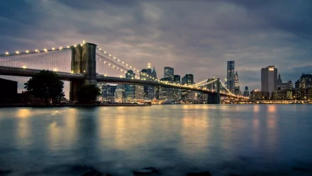 HD desktop wallpaper featuring the illuminated Brooklyn Bridge spanning over water with the Manhattan skyline in New York City under a moody evening sky.