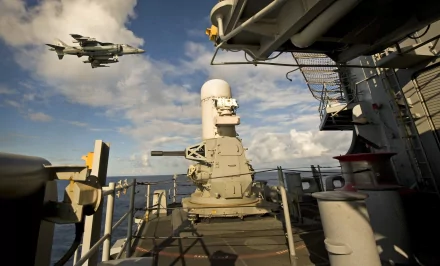 View from the deck of the USS Bonhomme Richard (LHD 6) amphibious assault ship with a McDonnell Douglas AV-8B Harrier II jet flying overhead against a partly cloudy sky.