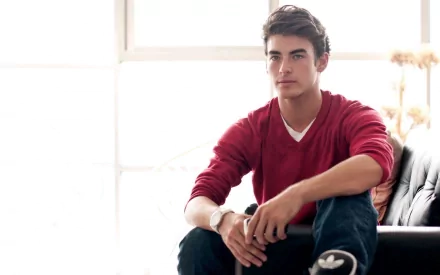 A man wearing a red shirt sits on a couch in a brightly lit room. The image is a beautiful HD desktop wallpaper and background.